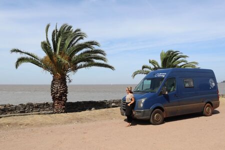 Ein blauer Van in Colonia del Sacramento an der Promenade stehend. Zwei Palmen im Hintergrund, sowie der gräuliche Rio de la Plata. Eine junge Frau lehnt vorne am Van und genießt die Aussicht bei leicht bewölktem und trotzdem blau-hellem Himmel.