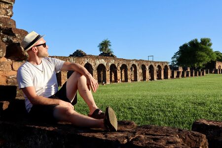 Ein Reisender sitzt auf alten Steinen und schaut in die Ferne. Hinter ihm die alte Jesuitenmission in Trinidad, Paraguay. Der Himmel ist blau, die Wiese ist grün und das Bild strahlt aus, den Moment aufzusaugen.