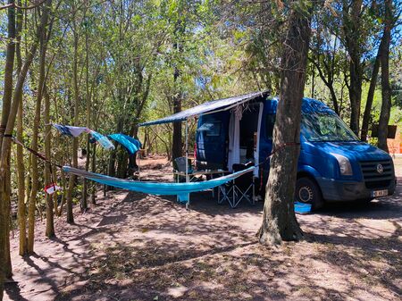 Ein blauer Campervan auf einem Campingplatz in Uruguay. Er ist umgeben von Bäumen, eine blaue Hängematte hängt im Vordergrund und im Hintergrund stehen Tisch und Stühle unter der ausgefahrenen Markise. Ein Bild, das Lust auf Vanlife macht!