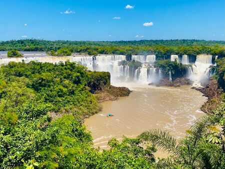 Ein weitläufiger Panoramablick auf die beeindruckenden Iguazú-Wasserfälle, umgeben von üppigem grünen Dschungel und blauem Himmel, mit dem Fluss im Vordergrund.