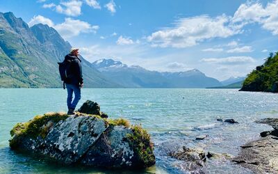 Ein Reisender steht auf einem Fels in einem türkisblauen See und schaut in die Ferne. Umliegend ein atemberaubendes Bergpanorama. Aufgenommen ist das Foto im Parque Nacional Tierra del Fuego.