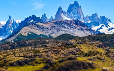 Eine atemberaubende Landschaft, vom Mirador de las Águilas aus fotografiert, die den Fitz Roy und den Cerro Torre bei klarem Wetter zeigt. Untermalt wird das Foto von satten Farben wie grün, blau und ein leichtes grau-violett, welches die Bergkette betont