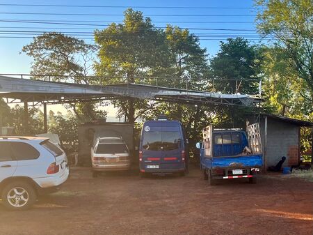 Unser blauer Van steht zwischen anderen Fahrzeugen auf dem Gelände einer Werkstatt in Paraguay, umgeben von Bäumen und unter einem klaren, blauen Himmel.