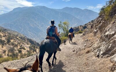 Reiter folgen einem schmalen Pfad in den Anden nahe Santiago de Chile. Die Szene zeigt eine Gruppe auf Pferden, eingebettet in eine trockene, bergige Landschaft mit Blick auf hohe Berge im Hintergrund.