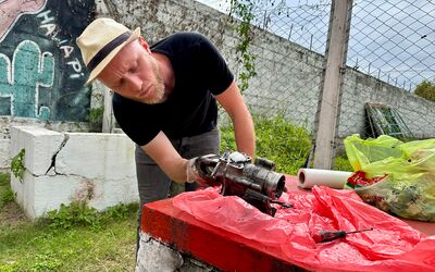 Ein Mann mit Hut arbeitet im Freien an einem Fahrzeugteil, das auf einer roten Unterlage liegt, während im Hintergrund eine Mauer mit Graffiti und ein Zaun zu sehen sind.